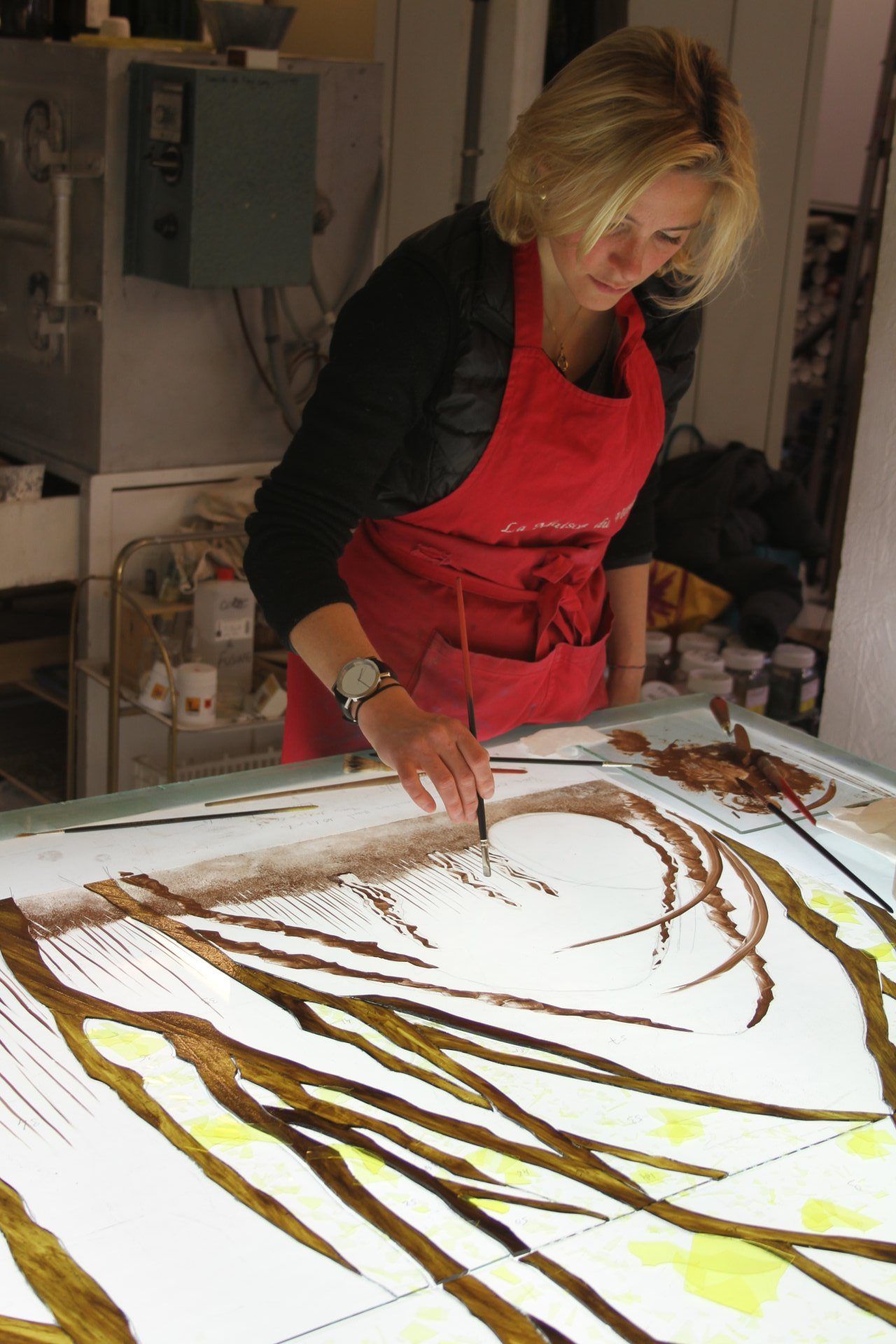 Femme en tablier rouge peignant une grande œuvre d'art illuminée à l'aide d'un pinceau fin. Atelier.