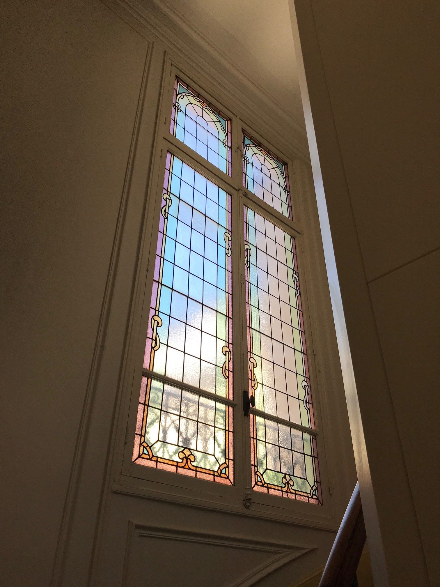 Haute fenêtre rectangulaire à vitraux, encastrée dans un mur blanc laissant apparaître un escalier.