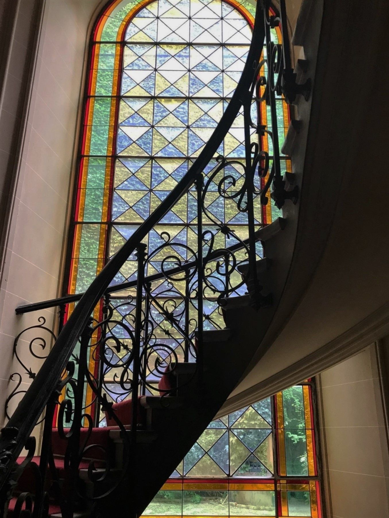 Escalier avec rampe en fer forgé ouvragée, adossé à une grande fenêtre en vitrail aux motifs géométriques bleus, jaunes et rouges.
