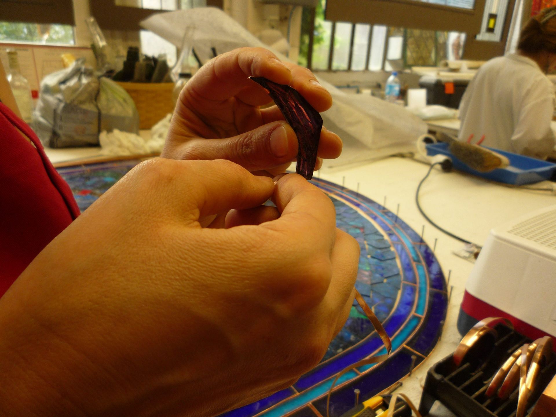 Des mains travaillent la feuille de cuivre sur un vitrail. Un panneau bleu et or se trouve en arrière-plan.