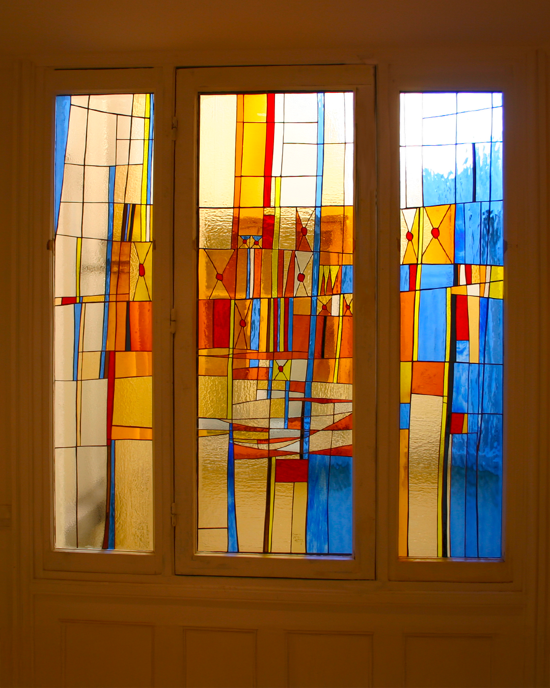 Vitrail à cadre blanc ; motif abstrait composé de blocs jaunes, oranges, rouges et bleus.