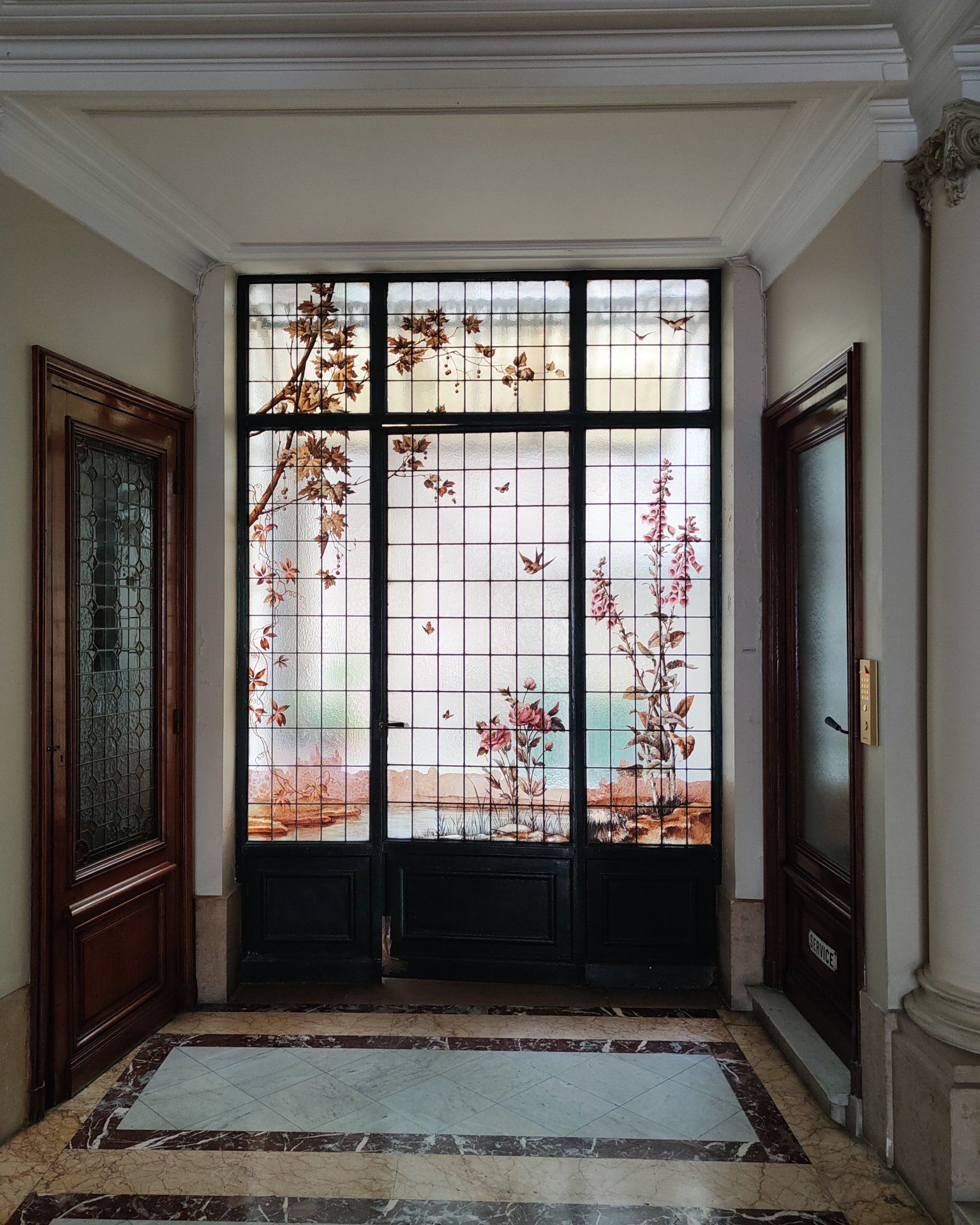 Couloir orné d'un vitrail, de portes en bois foncé et de moulures travaillées. Motifs floraux automnaux dans le verre.