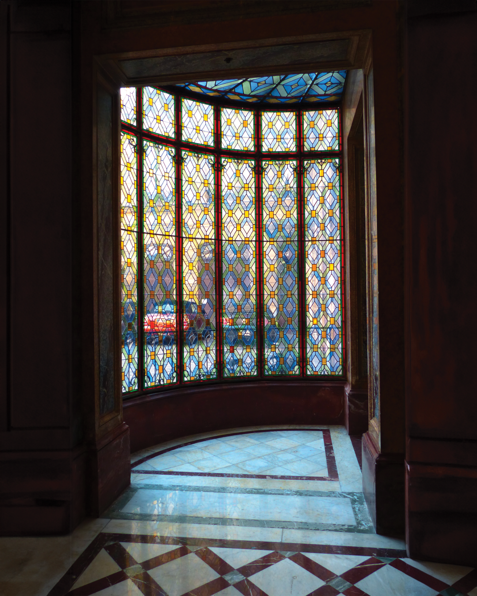 Vitrail dans une porte en bois. La lumière du soleil inonde l'intérieur. Une voiture rouge est visible à travers la vitre.