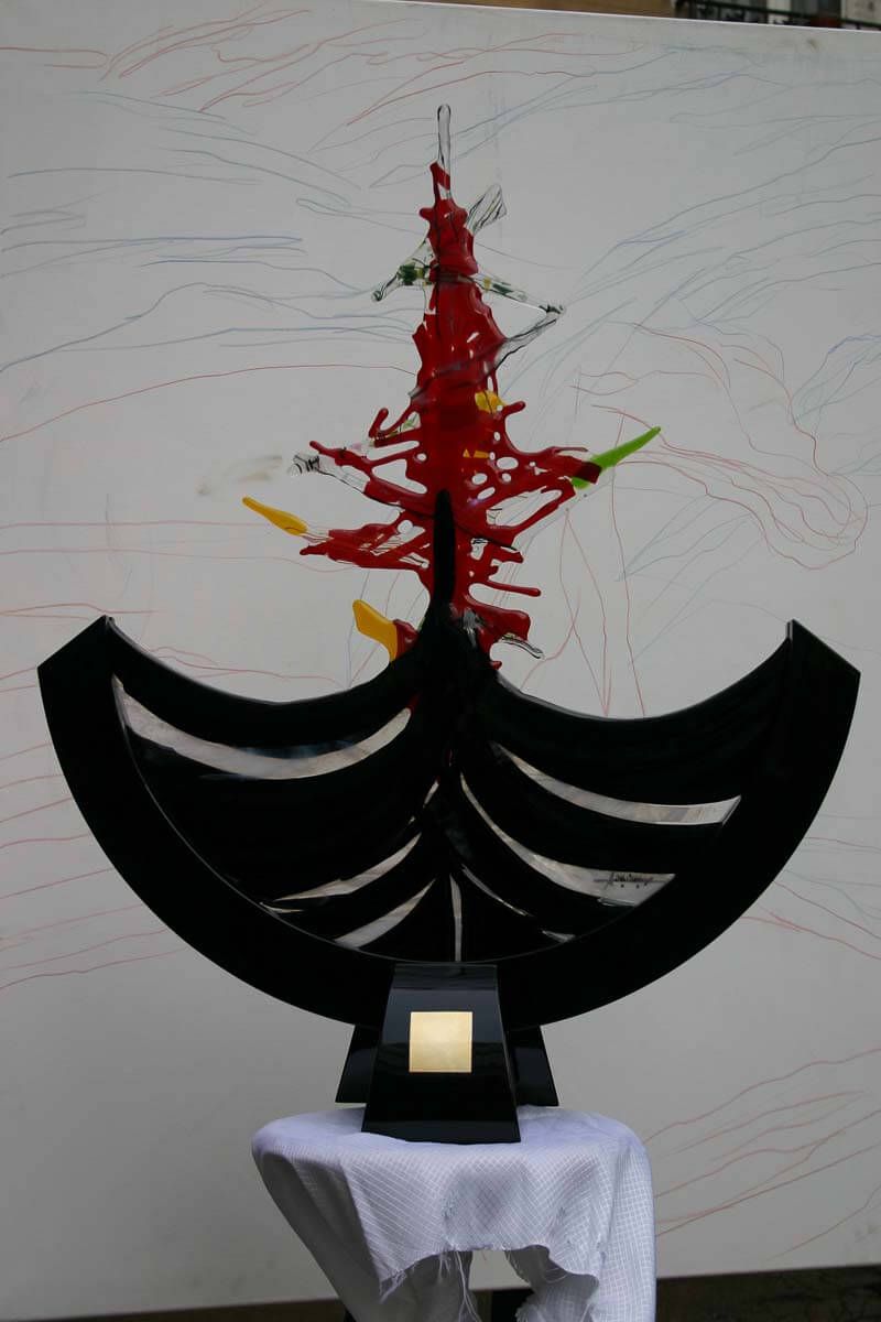 Sculpture noire ressemblant à un bateau, peinte en rouge, jaune et vert sur fond blanc.