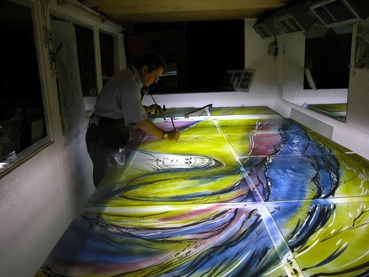 Une personne peint une grande fresque abstraite et colorée à l'intérieur d'une pièce. Le jaune, le bleu et le vert sont des couleurs dominantes.