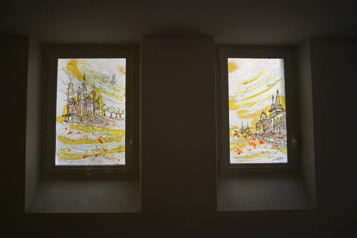 Deux fenêtres ornées de peintures de paysages urbains, principalement jaunes et brunes, encastrées dans un mur sombre.