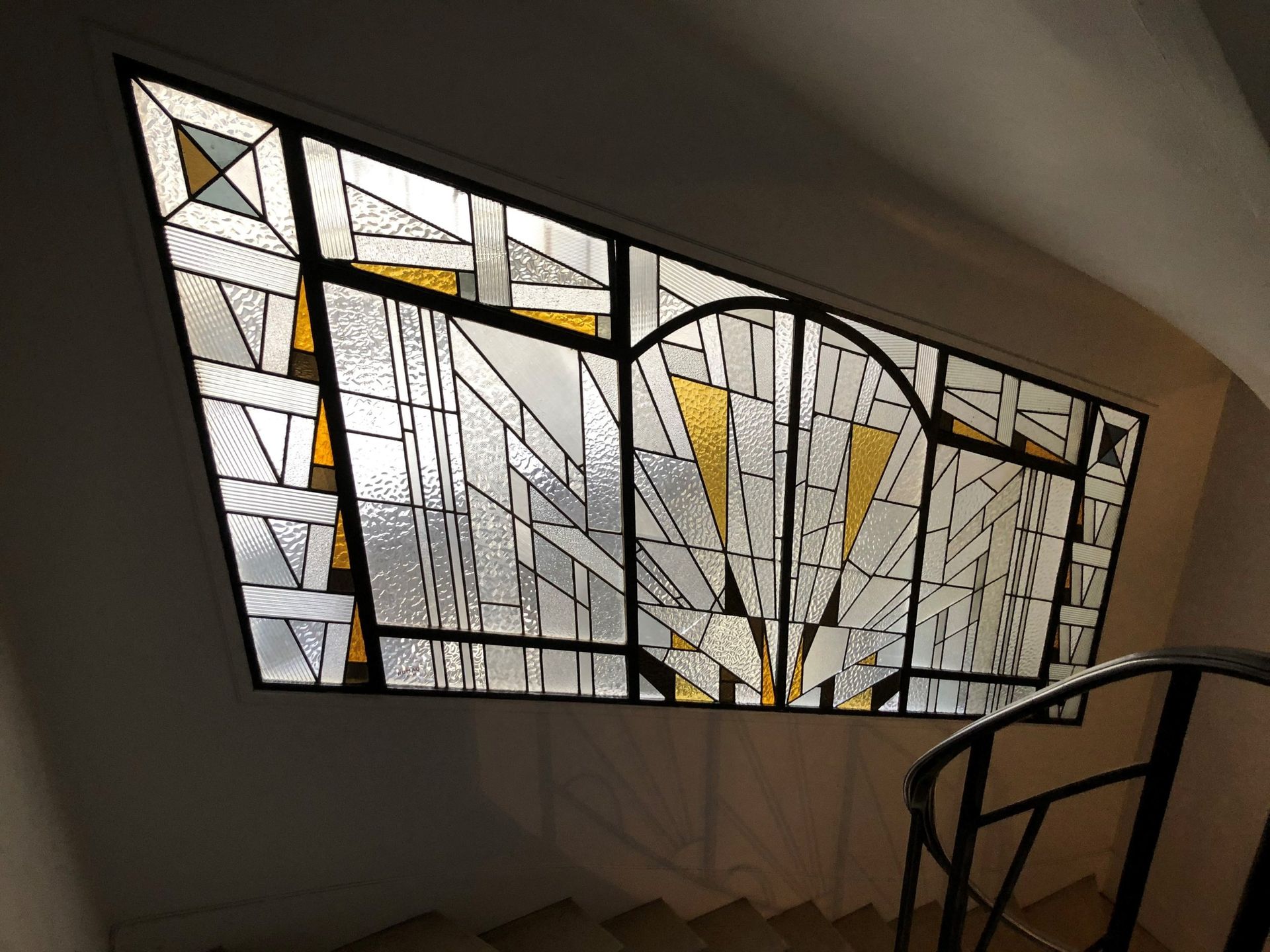 Vitrail à motifs géométriques, au-dessus d'un escalier à rampe noire et murs blanc cassé.