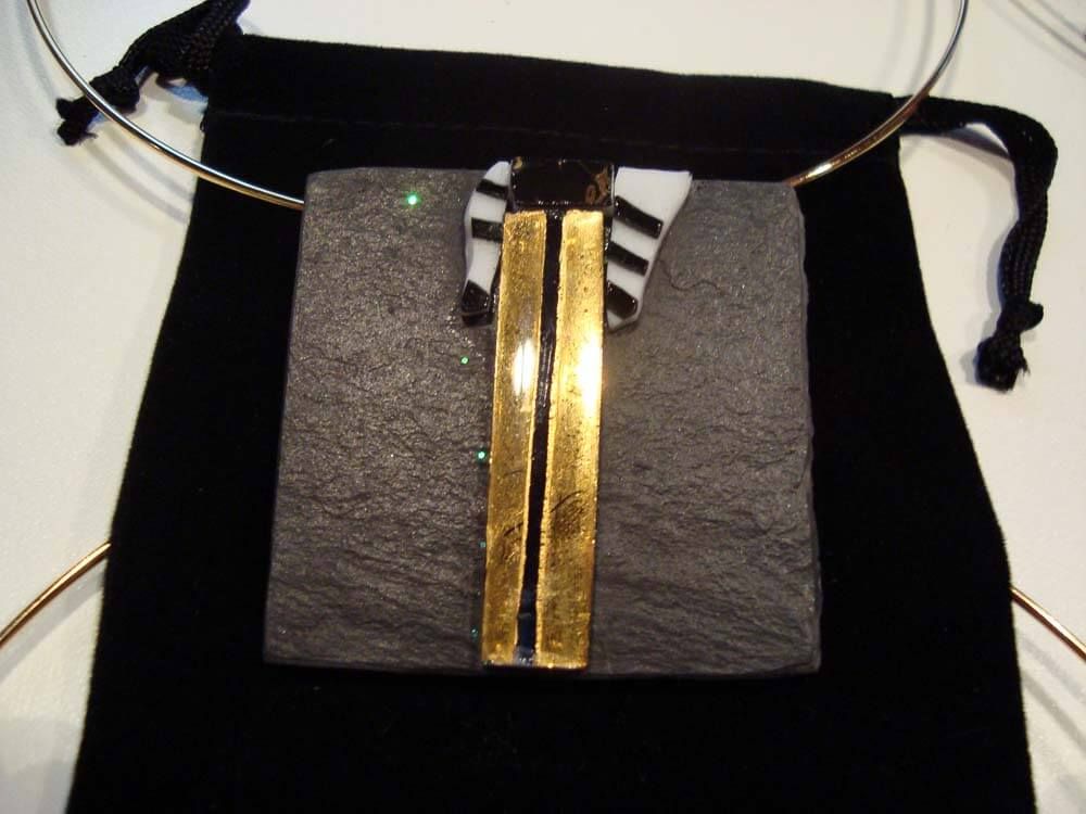 Collier avec pendentif carré en pierre grise, barres verticales dorées, accents de verre noir et blanc, sur fond noir.