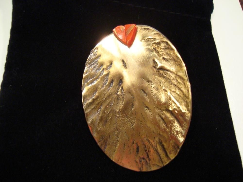 Broche ovale texturée de couleur or, ornée d'un petit accent rouge-orangé sur le dessus, posée sur une surface en velours noir.