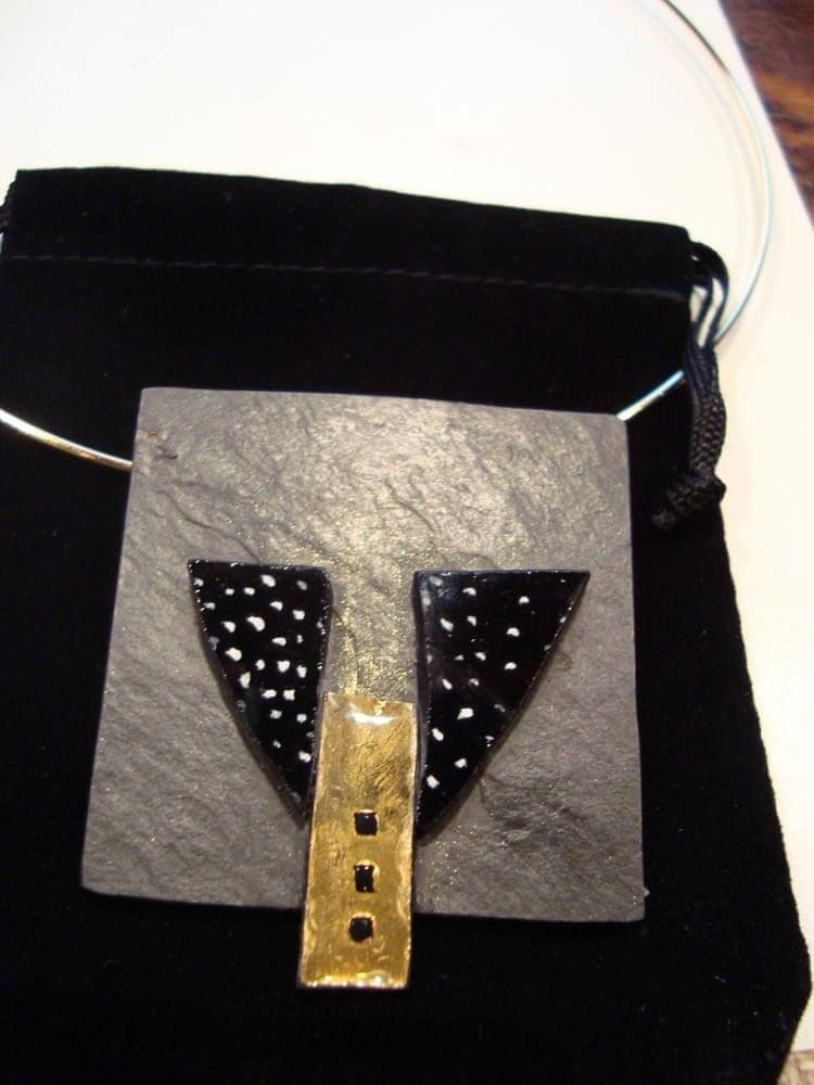 Collier pendentif carré en ardoise avec motif géométrique noir et or, sur pochette en velours noir.