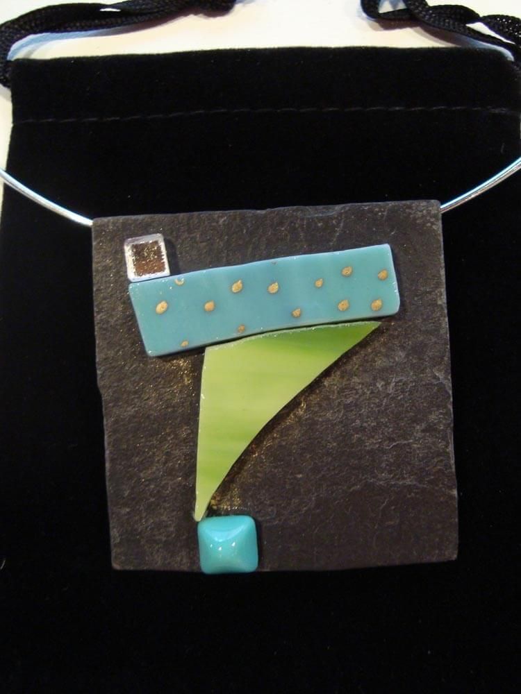 Collier pendentif abstrait avec des accents turquoise, verts et dorés sur un fond carré sombre.