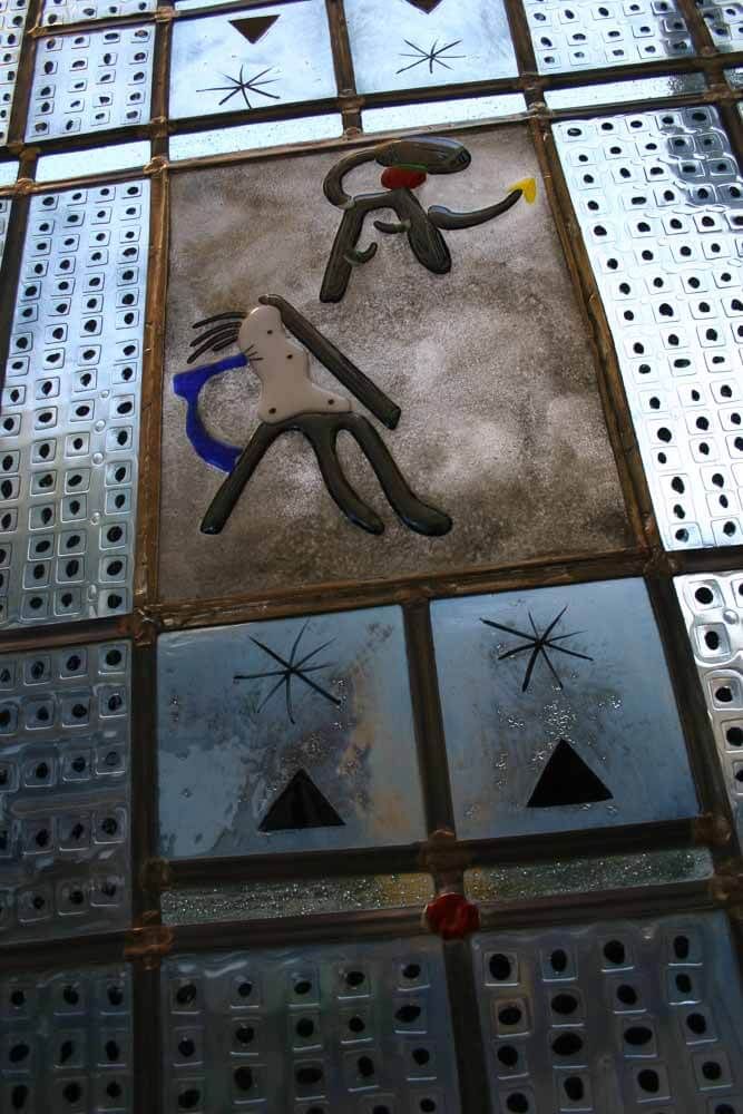 Panneau de vitrail orné de deux figures stylisées, d'étoiles, de triangles et de carrés à motifs dans des tons métalliques et bleus.