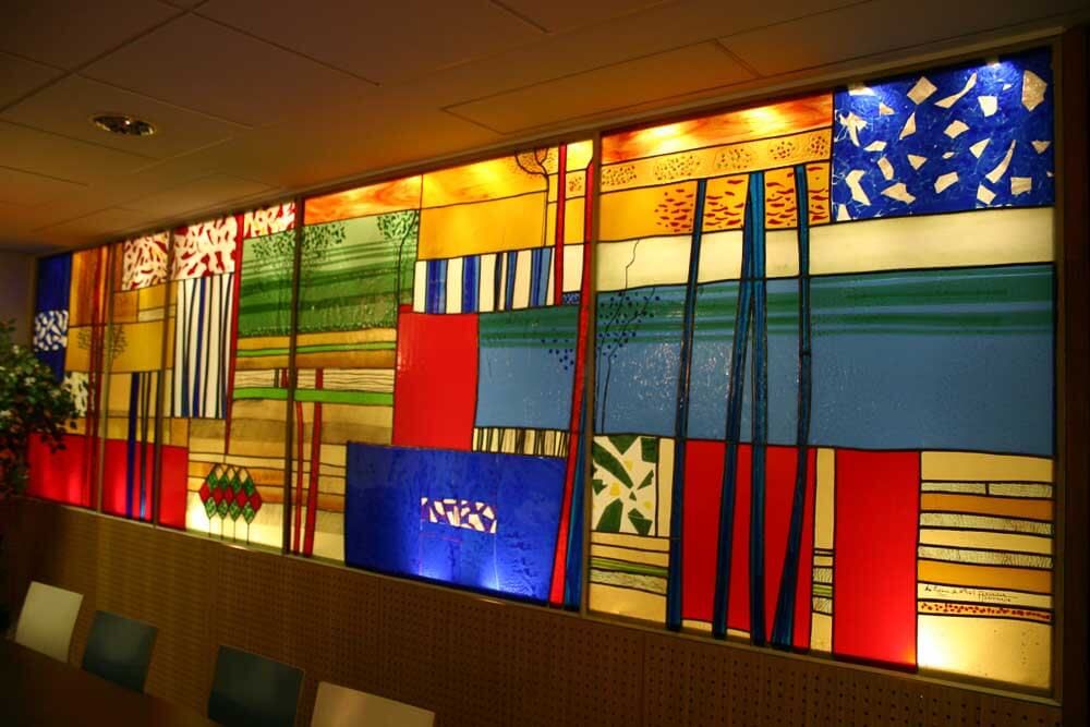 Vitrail mural composé de panneaux rectangulaires de couleurs et de motifs variés, illuminé.
