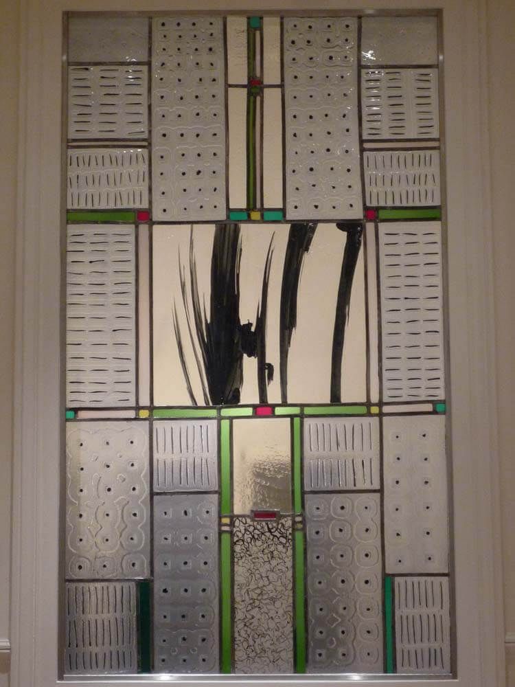 Vitrail au motif abstrait. Coups de pinceau noirs sur fond crème, entourés de panneaux à motifs avec des accents verts et rouges.