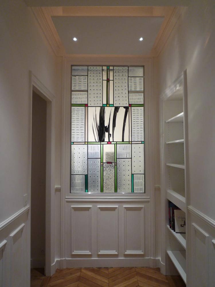 Couloir aux murs blancs, étagères intégrées, et un panneau en vitrail orné d'art abstrait, avec des boiseries blanches en dessous.