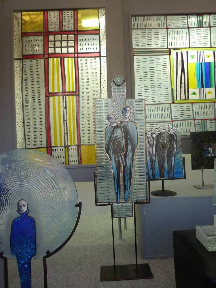 Exposition d'œuvres d'art : vitraux, figures humaines en verre, sphère et formes urbaines, dans les tons jaunes, bleus et blancs.