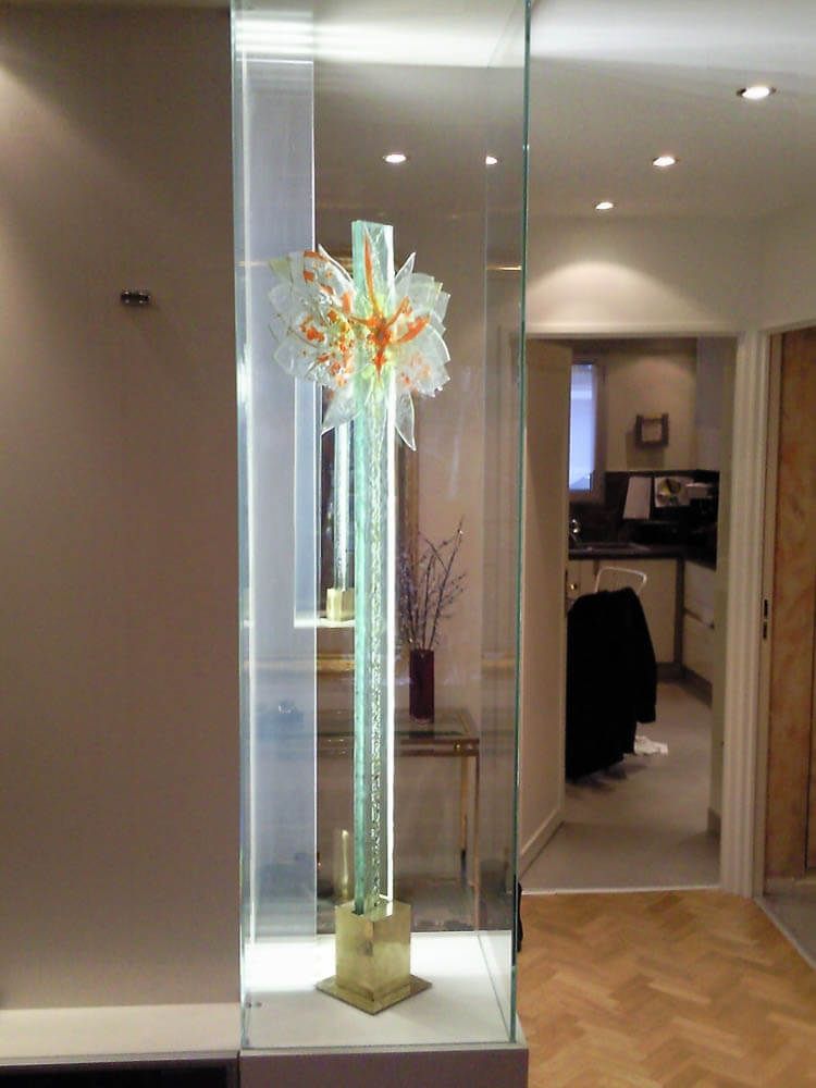 Sculpture en verre dans une vitrine éclairée ; verte et dorée avec des touches d’orange. Vue sur une cuisine et un couloir.