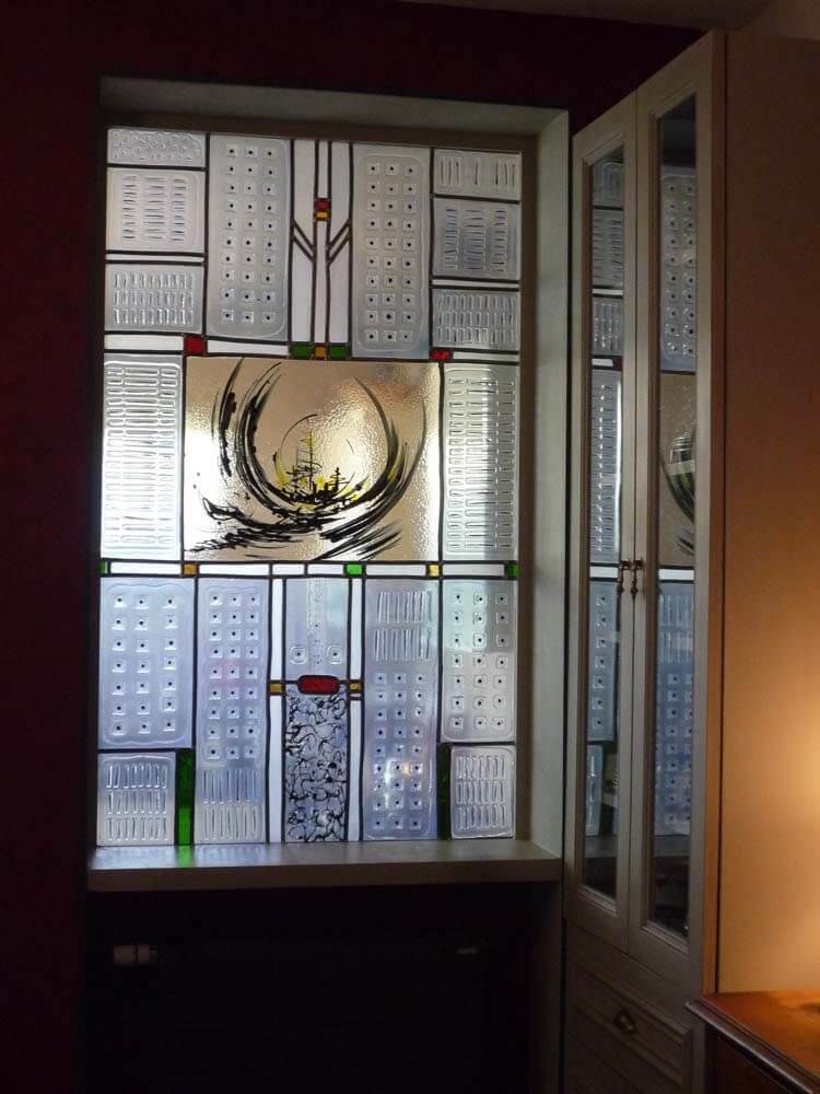 Vitrail à motifs abstraits blancs, noirs, verts et jaunes, dans un cadre blanc, avec une porte de placard à droite.