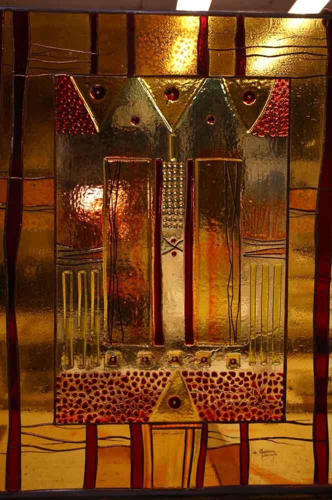 Œuvre en vitrail ornée de formes géométriques dorées et rouges, illuminée par l'arrière.