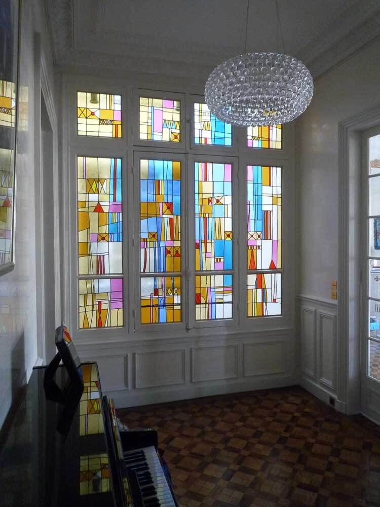Un vitrail aux motifs géométriques colorés orne un intérieur lumineux ; un piano est placé en dessous et un lustre est suspendu au-dessus.