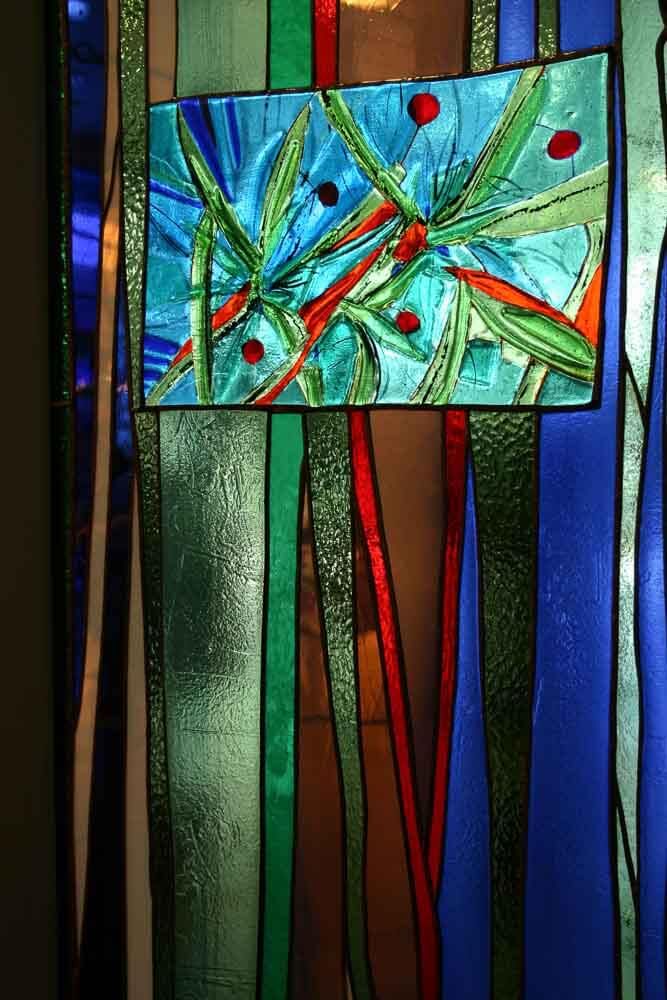 Vitrail à décor abstrait. Fond bleu avec des accents verticaux verts, rouges et bruns. Le panneau central représente une scène botanique.