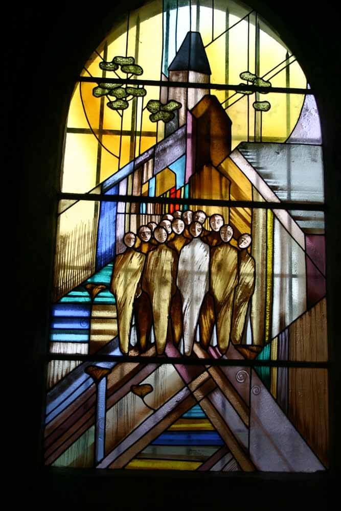 Vitrail représentant des figures géométriques humaines devant un paysage urbain architectural ; couleurs chaudes.