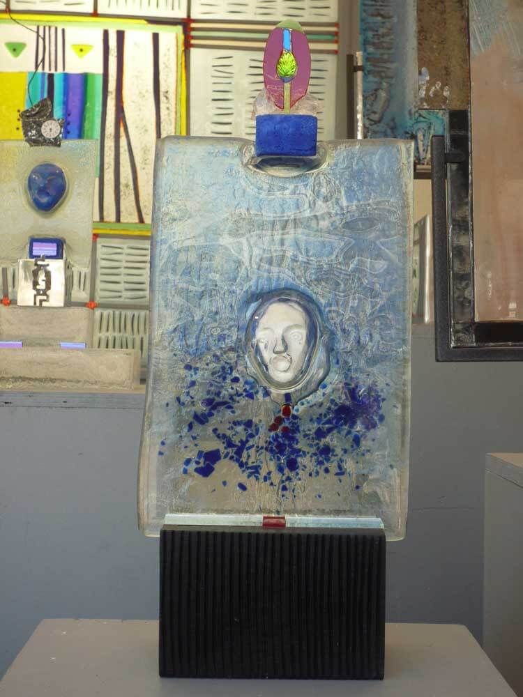 Sculpture abstraite au visage peint en bleu, montée sur un socle noir. Des touches de violet, de bleu et de blanc y sont intégrées.