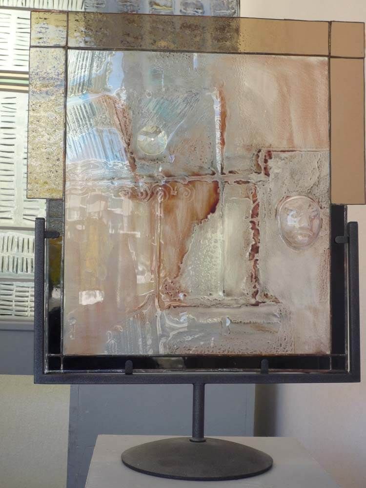 Œuvre d'art abstraite en verre sur support métallique, présentant des textures superposées et des tons terreux de brun, de beige et de bleu.