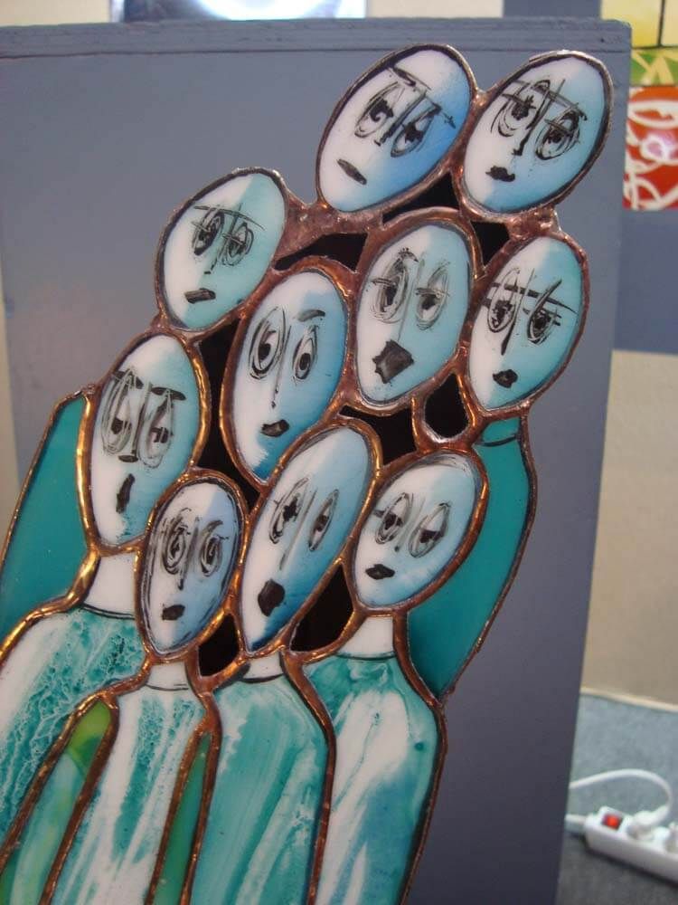 Panneau de vitrail : groupe de visages bleus et allongés à l’expression surprise. Contours cuivrés et figures turquoise.
