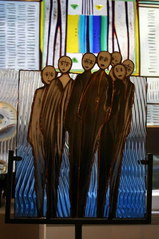 Sculpture en verre représentant sept personnages aux tons neutres, se détachant sur un fond bleu texturé.