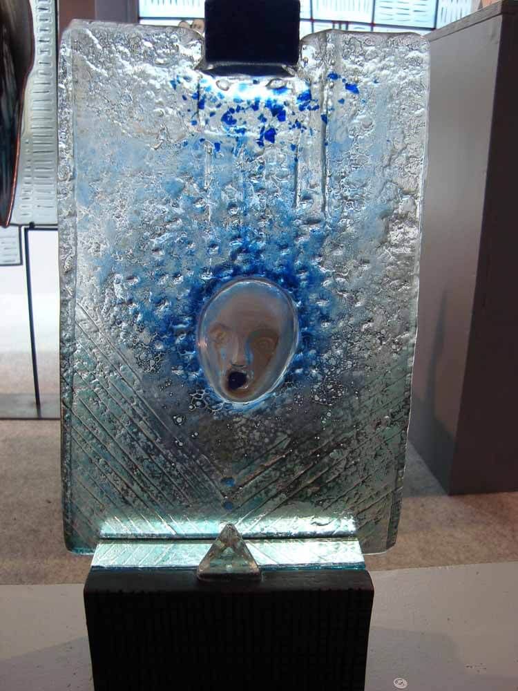 Sculpture : Panneau de verre rectangulaire orné de touches de bleu et d’un visage. Un prisme repose sur un socle noir.