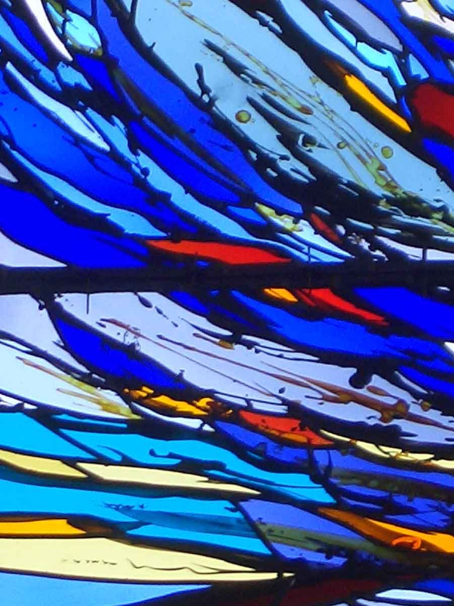 Vitrail abstrait aux formes fluides bleues, rouges et jaunes ; la lumière visible le traverse.