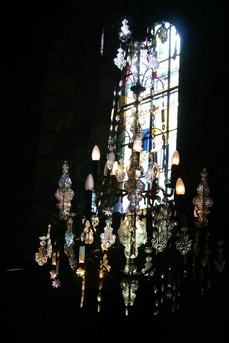 Un lustre orné dans une pièce faiblement éclairée, avec un vitrail en arrière-plan, illuminé par la lumière du soleil.