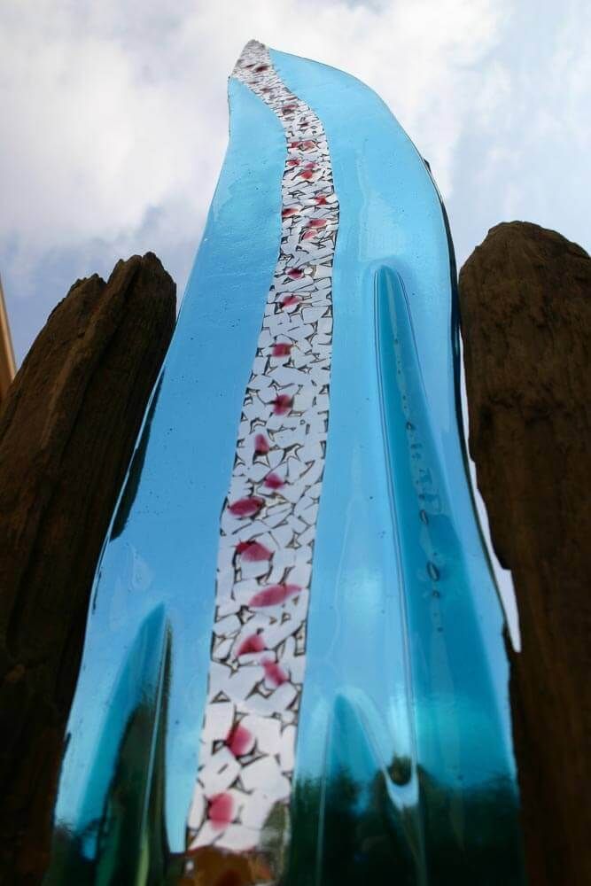 Sculpture en verre bleu ornée d'une rayure blanche et rose, flanquée de bois brun sur fond de ciel.