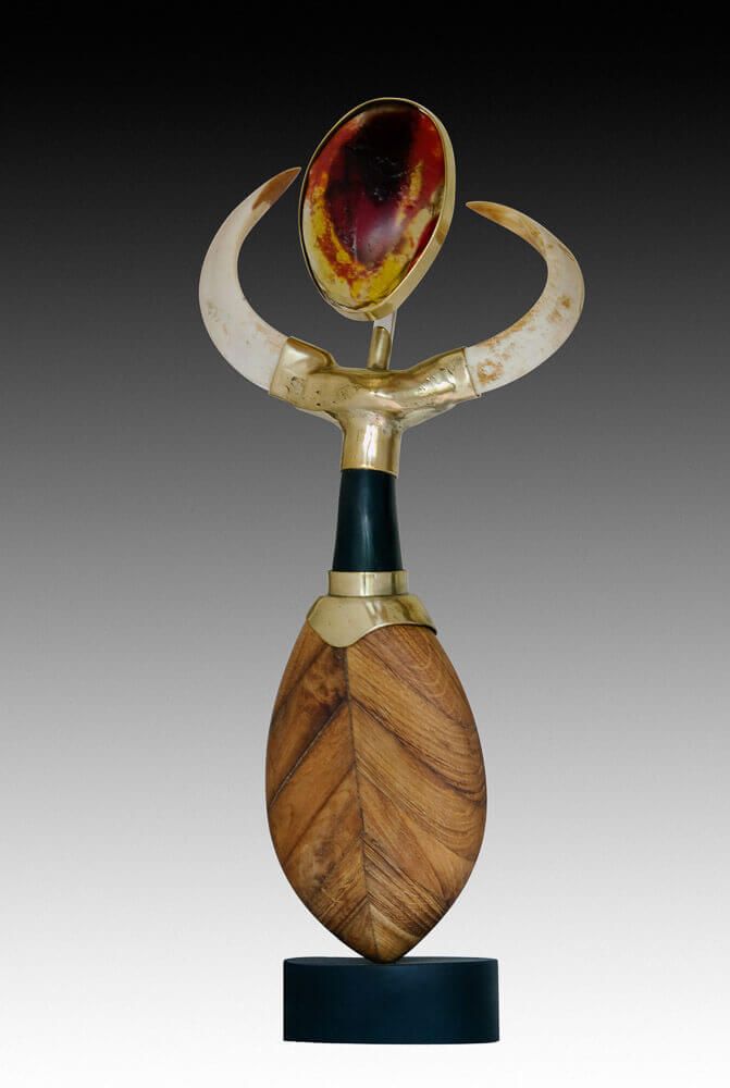 Sculpture abstraite représentant une feuille de bois sur un socle noir. Des bras dorés s'élèvent d'une colonne sombre soutenant une gemme ovale.