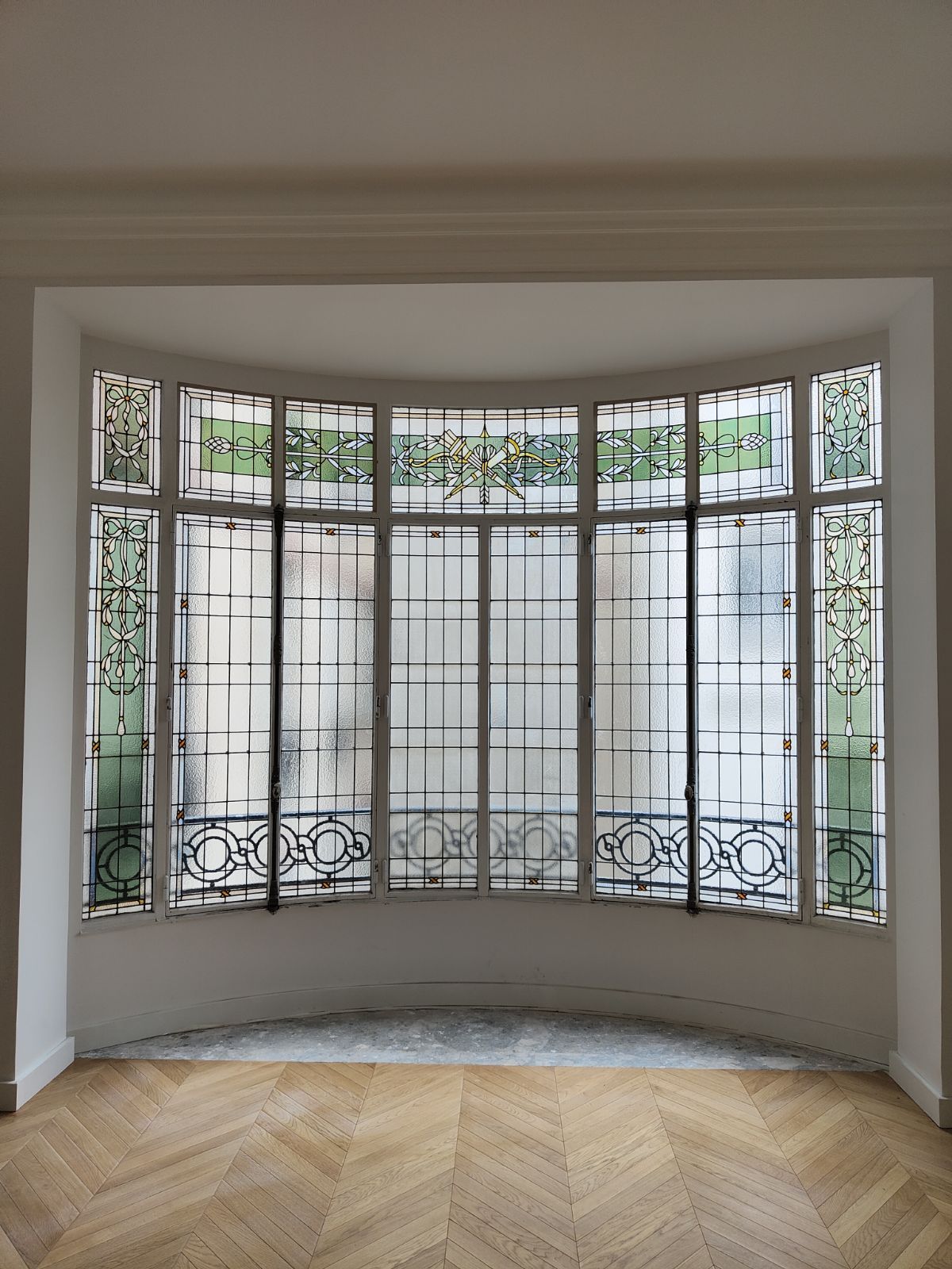 Baie vitrée cintrée ornée de détails en métal noir. La lumière filtre à travers le verre dépoli. Parquet à chevrons.