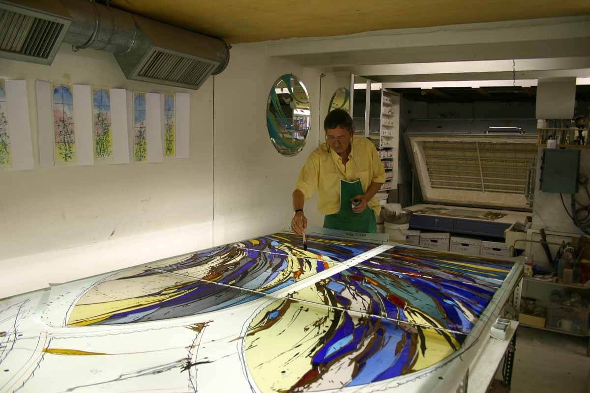 Artiste travaillant sur un panneau de vitrail ; dans son atelier, il peint un motif coloré sur du verre.