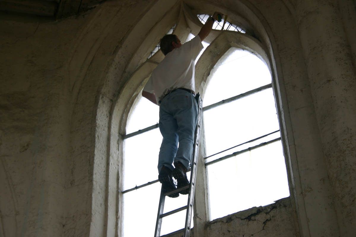 Une personne sur une échelle, travaillant sur une haute fenêtre cintrée à l'intérieur d'un bâtiment.