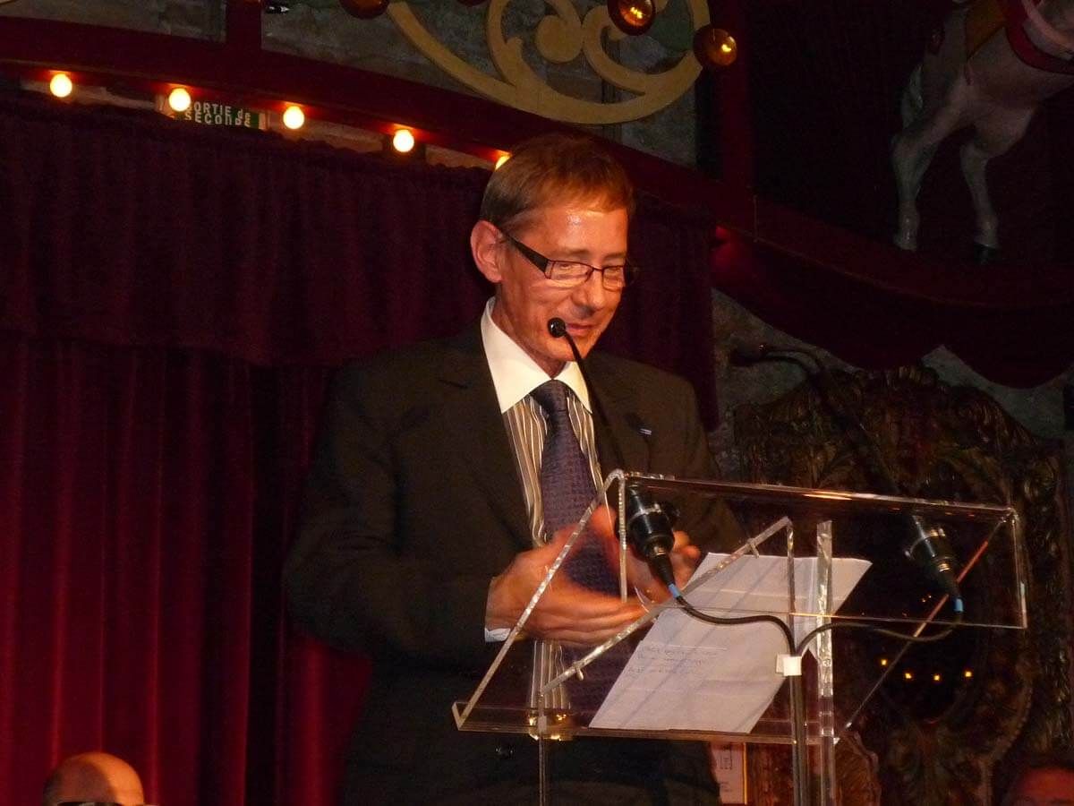 Un homme en costume prend la parole à la tribune, souriant, sur fond de rideau rouge.