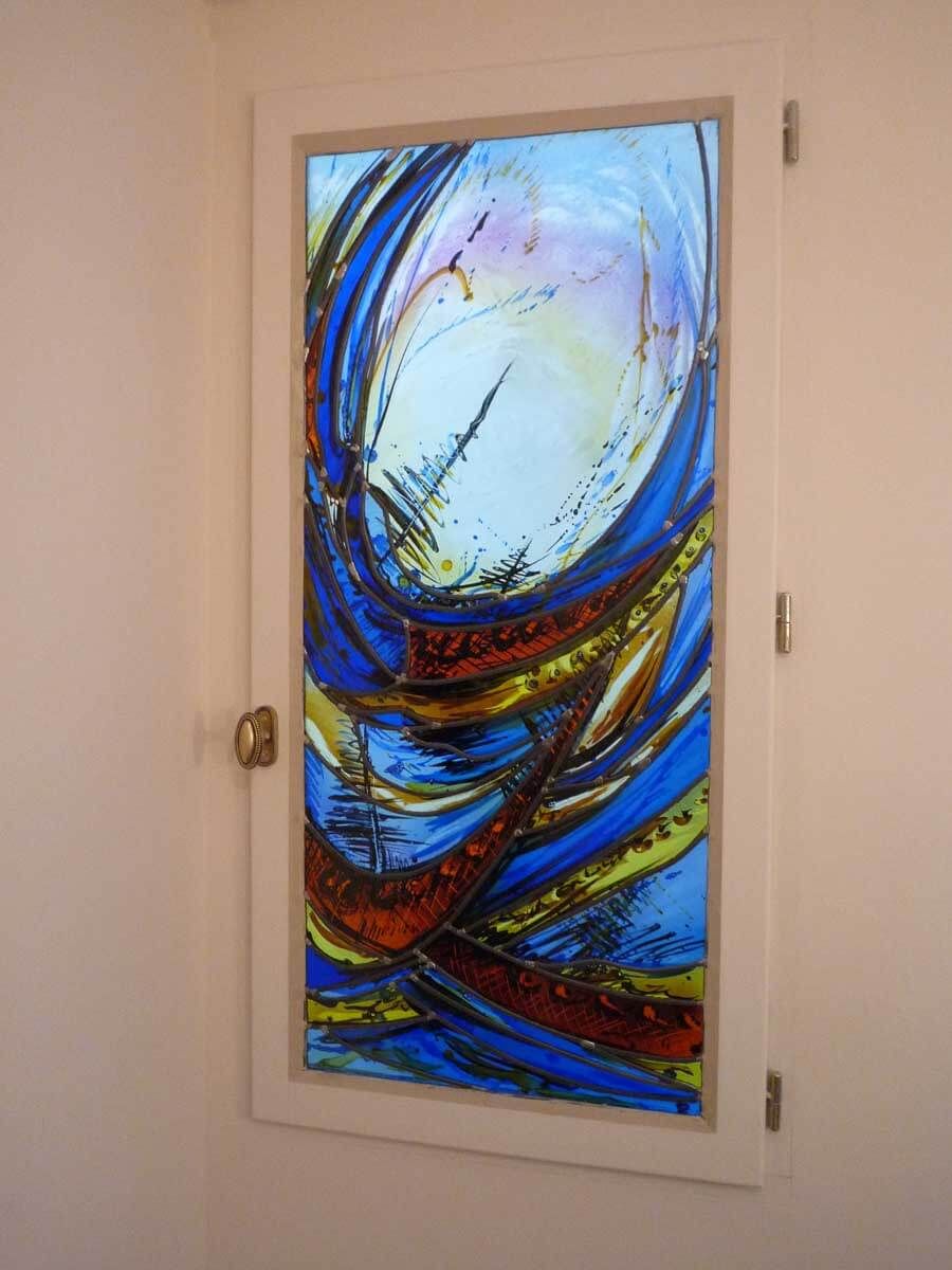 Vitrail à motif abstrait bleu, orange et jaune, encadré de blanc et posé sur un mur beige.