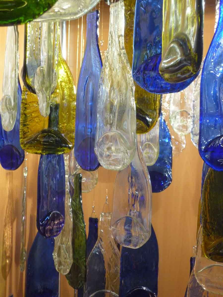 Œuvre d'art composée de vitraux en formes de bouteilles en verre suspendues de différentes couleurs, notamment bleues, jaunes et transparentes.