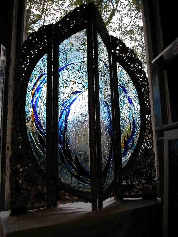 Vitrail orné de volutes bleues et de barres verticales, encadré d'un cadre sombre et ouvragé, installé dans une fenêtre.