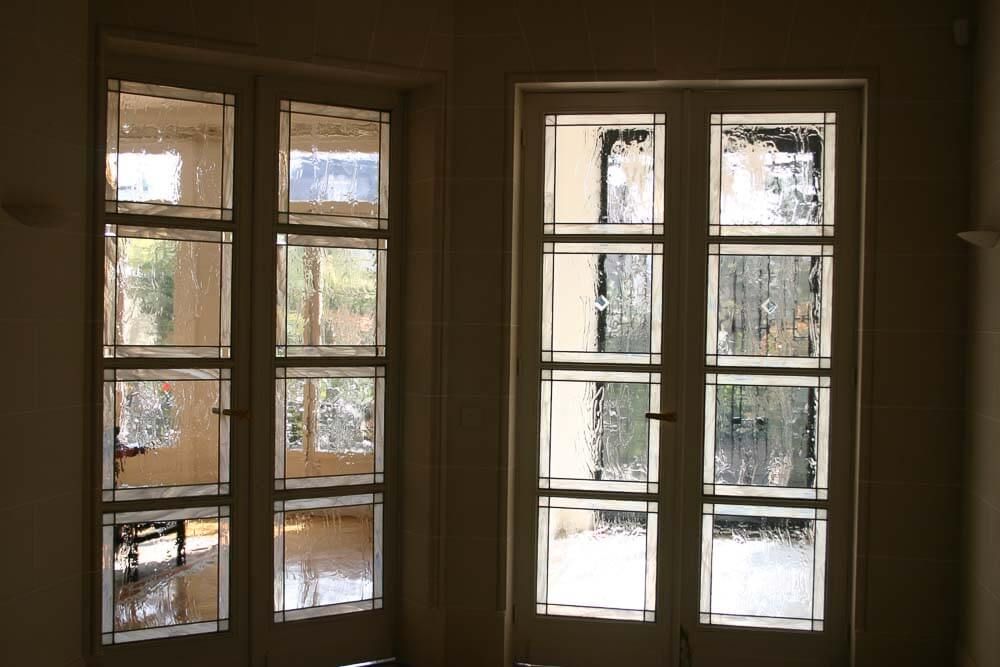 Deux hautes fenêtres à petits carreaux, recouvertes de verre dépoli, vues de l'intérieur d'une pièce. La lumière du soleil filtre à travers les vitres opaques.