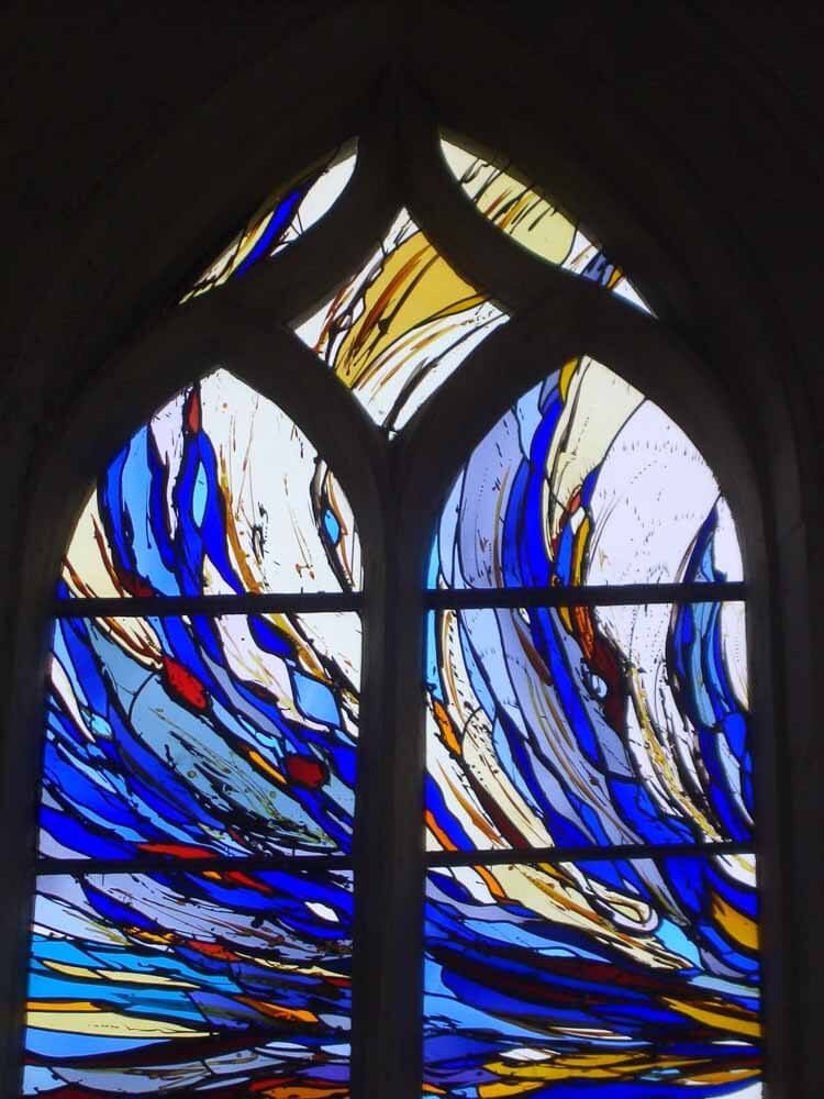 Vitrail orné de motifs abstraits tourbillonnants bleus, blancs et jaunes, enchâssé dans une arche en pierre.