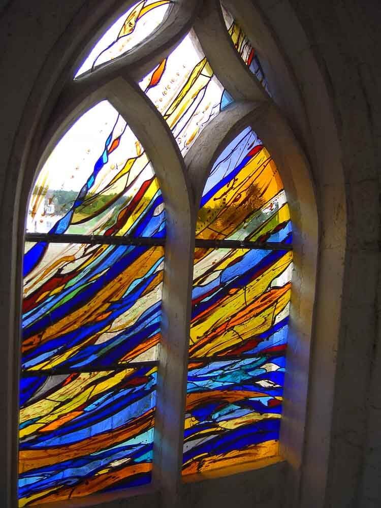 Vitrail abstrait coloré dans un cadre en pierre arqué ; bleus, jaunes, oranges et blanc.