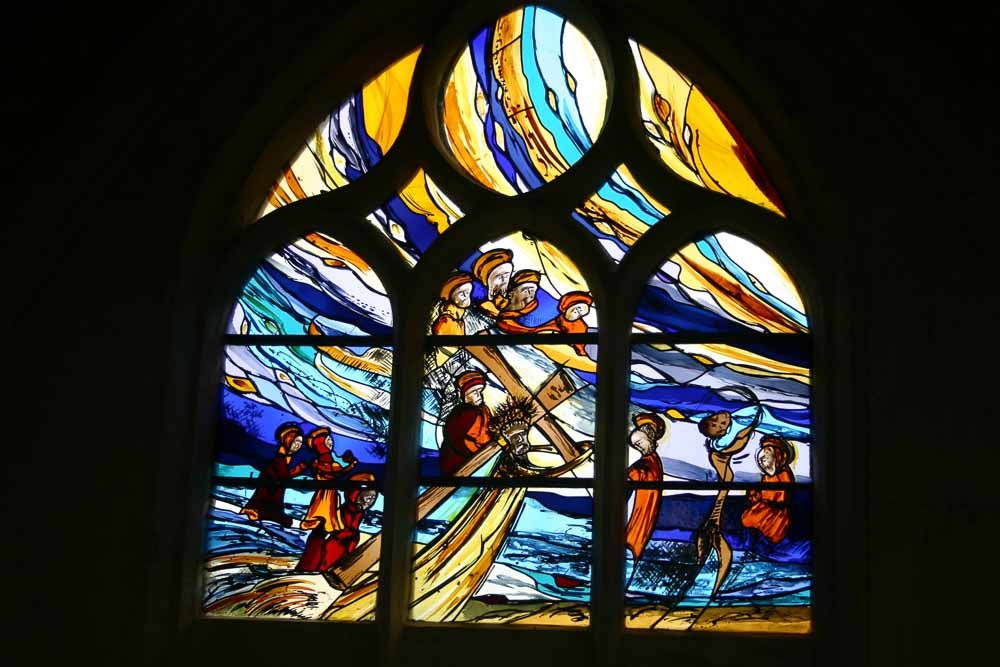 Vitrail représentant des personnages sur une barque au milieu de vagues bleues, portant une croix. Ciel jaune et bleu.