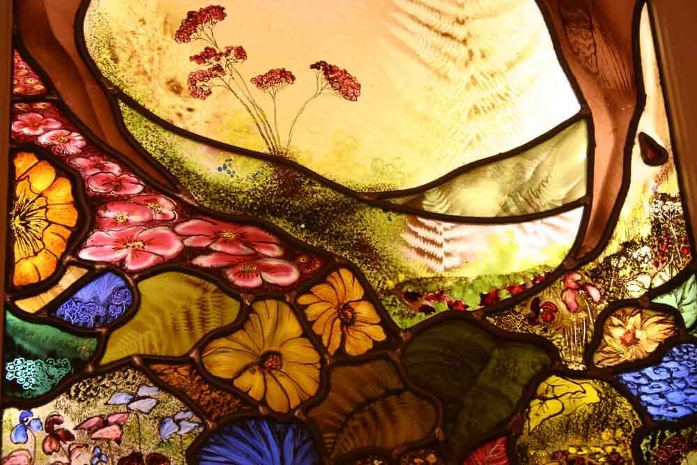 Vitrail : scène florale colorée avec des fleurs orange, roses, jaunes et bleues sur un fond clair.