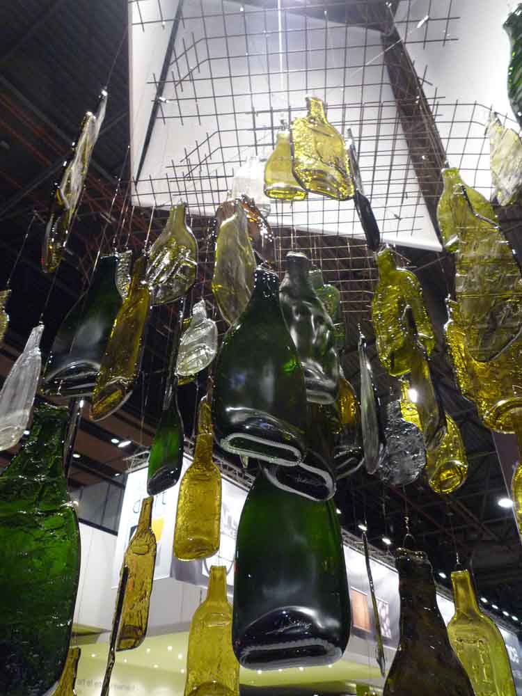 Installation au plafond composée de vitraux en formes de bouteilles de vin en verre et d'éclats de verre de couleurs variées, suspendues à une grille.