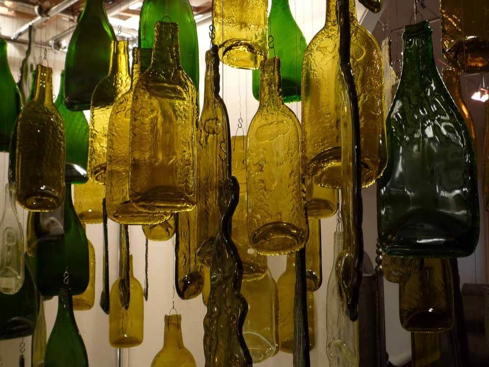 Sculpture suspendue composée de vitraux en formes de bouteilles en verre vert et ambré recyclées, à l'intérieur, éclairée par le haut.
