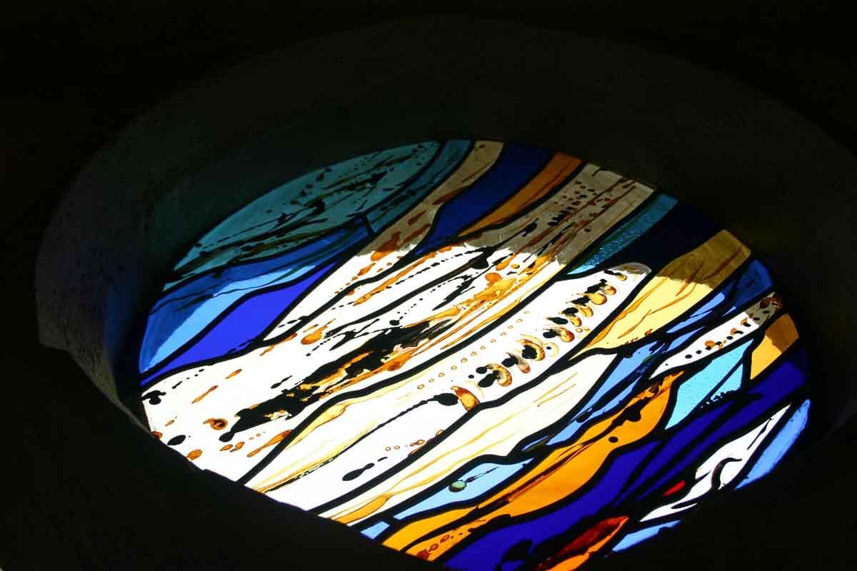 Vitrail circulaire à motifs ondulés bleus, jaunes et orange, encadré d'une pierre sombre.